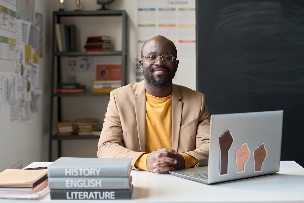 Portrait of African American teacher smiling at camera while sitting at table with laptop and books in the classroom