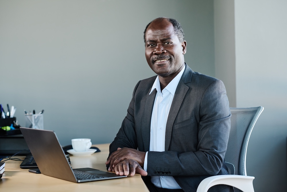 Happy experienced chief executive officer in formalwear looking at camera while sitting by workplace in office and networking